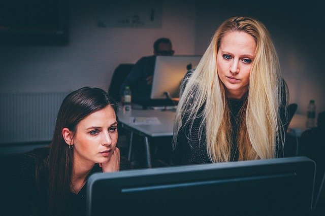 A brunette woman, sitting down, and a blonde woman, standing up, work together to solve challenges around Cisco Meraki Licensing co-termination challenges 